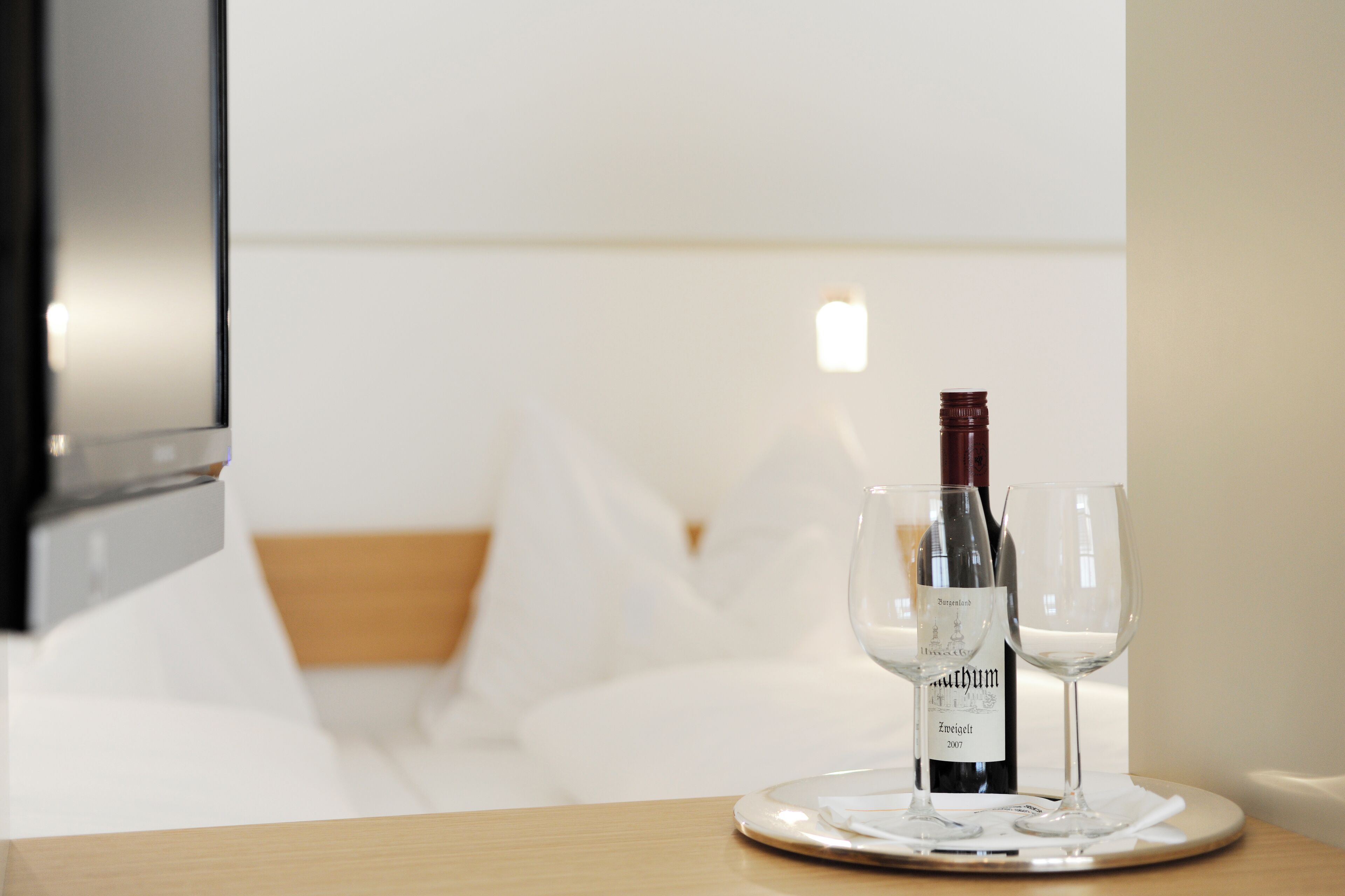 Habitación de hotel con cama, mesita de noche con una botella de vino y copas, y televisor instalado en la pared.