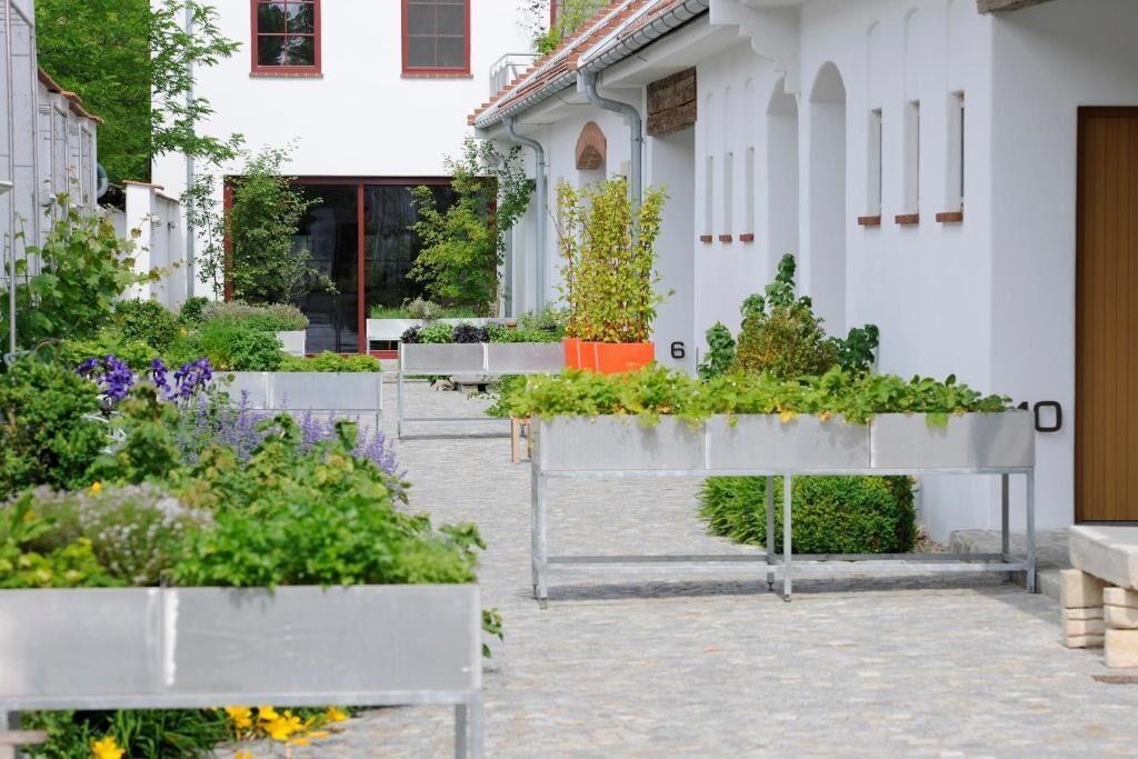 Ein Innenhof mit erhöhten Pflanzgefäßen, in denen verschiedene Pflanzen und Blumen stehen, einem gepflasterten Weg und einem weißen Gebäude mit Bogenfenstern und einer Holztür.