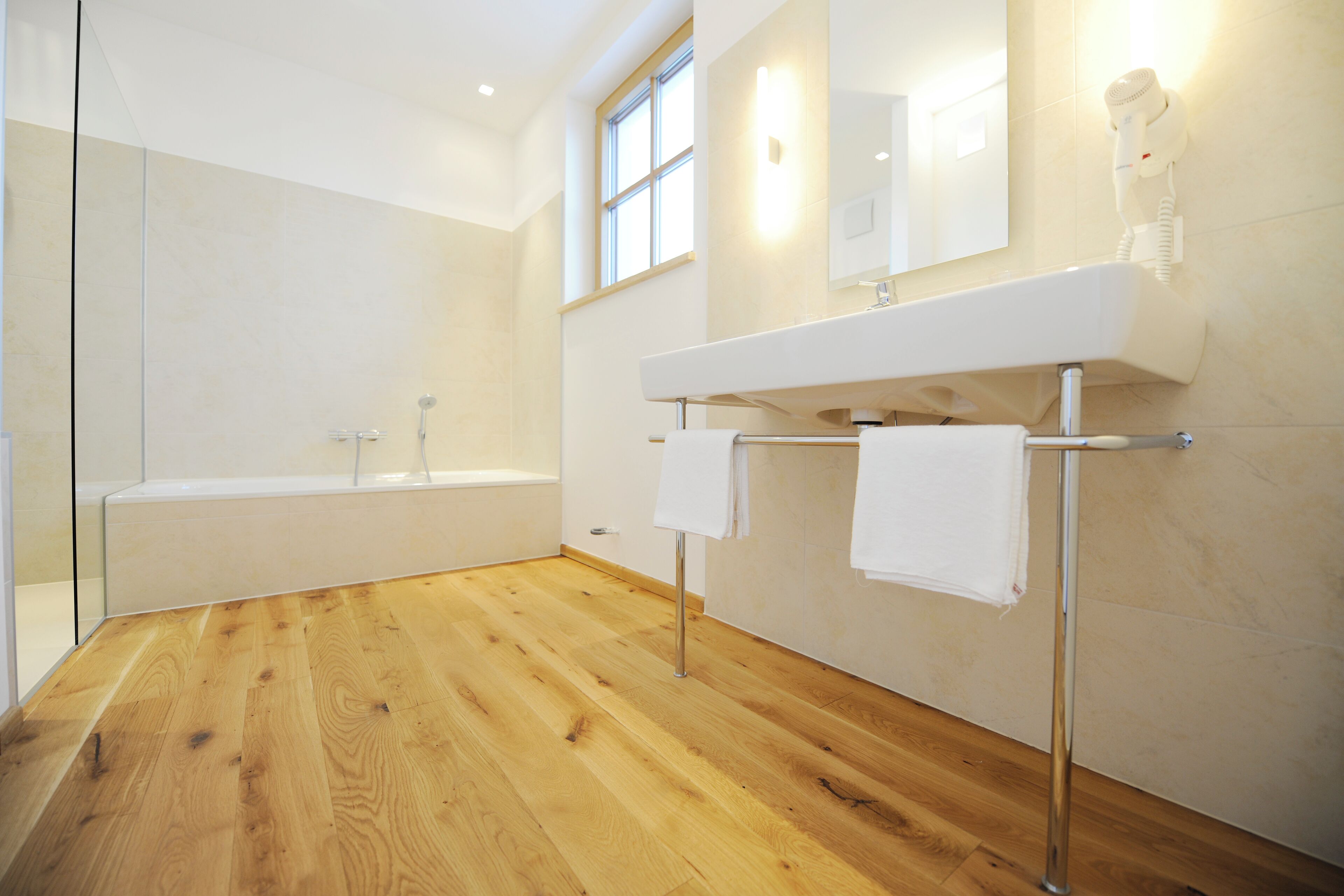 Ein modernes Badezimmer mit Holzboden, einer Badewanne, einem großen Spiegel und einem an der Wand montierten Fön.