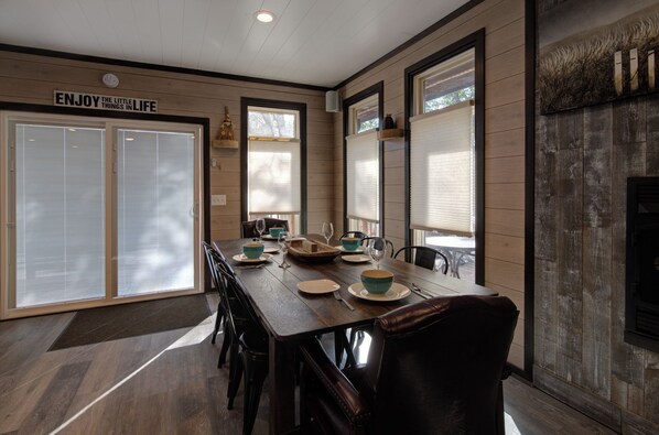 Un comedor con una mesa de madera dispuesta para cuatro personas, rodeada de sillas. A la derecha hay una chimenea y un cartel sobre la puerta que dice "ENJOY THE LITTLE THINGS IN LIFE".