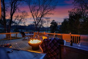 Una terraza con asientos exteriores, una fogata y vistas a árboles y al atardecer.