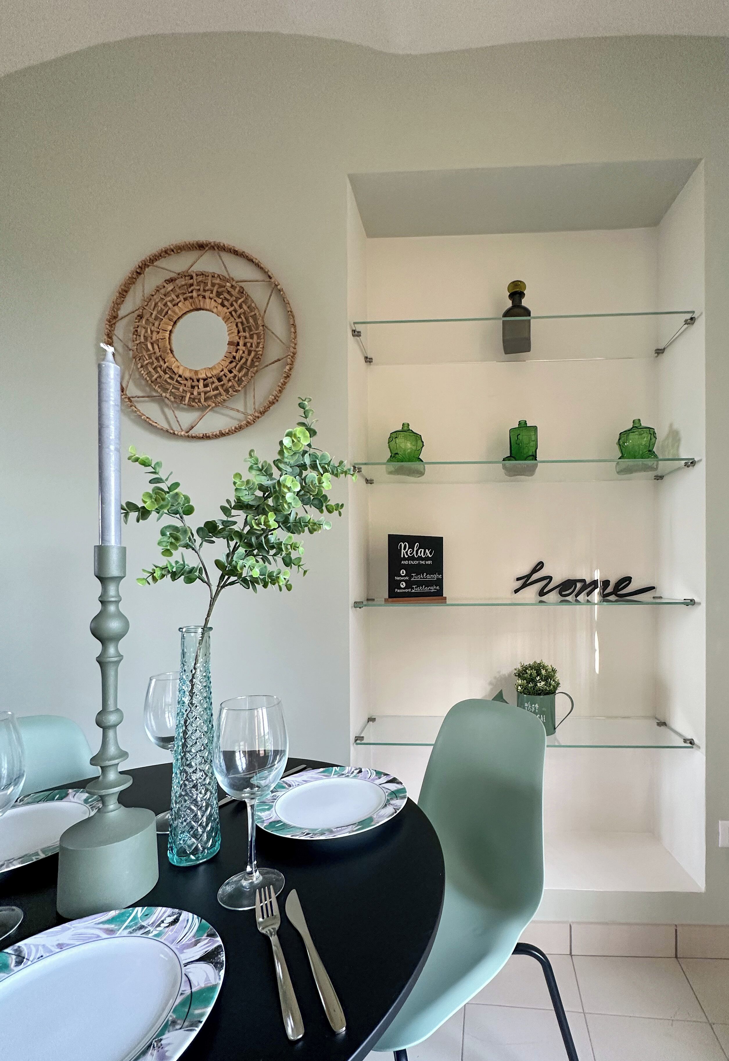 Un área de comedor con una mesa dispuesta para dos personas, un tapiz decorativo y una estantería con objetos decorativos.