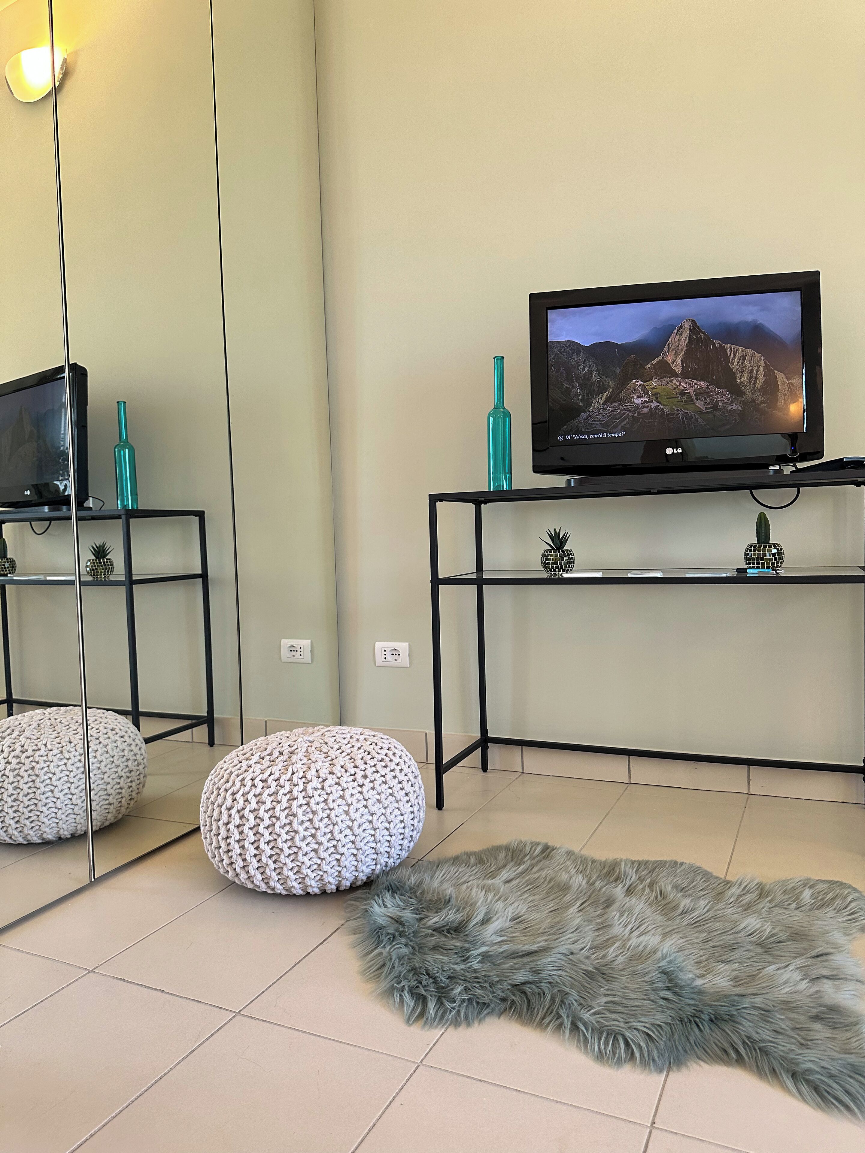 Ein Wohnzimmer mit Fernseher auf einem schwarzen Schrank, zwei Hocker, einem Fellteppich und einer Wandleuchte.