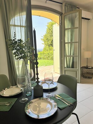 A dining table set with plates, glasses, cutlery, and a vase with greenery, positioned near a window with curtains, and an air conditioning unit on the wall. - Just Langhe Casa Dolcetto (Piemonte)