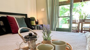 Frühstückswagen mit Teekanne, zwei Tassen und einer kleinen Pflanze auf einer gewebten Matte, platziert auf einem Bett mit Blick ins Grüne.