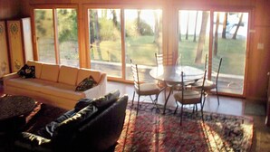 A living room with a sofa, a coffee table, and a dining table with chairs. Large windows provide a view of the outdoors.