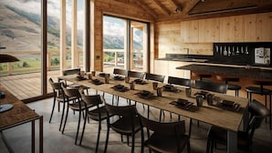A dining area with a long wooden table set for 12, surrounded by chairs, with a kitchen area and large windows offering a mountain view. - Chalet, wifi, sauna, terrace, fireplace or stove, parking, tv, ski locker, 250m², Les 2 Alpes (Les Deux Alpes)