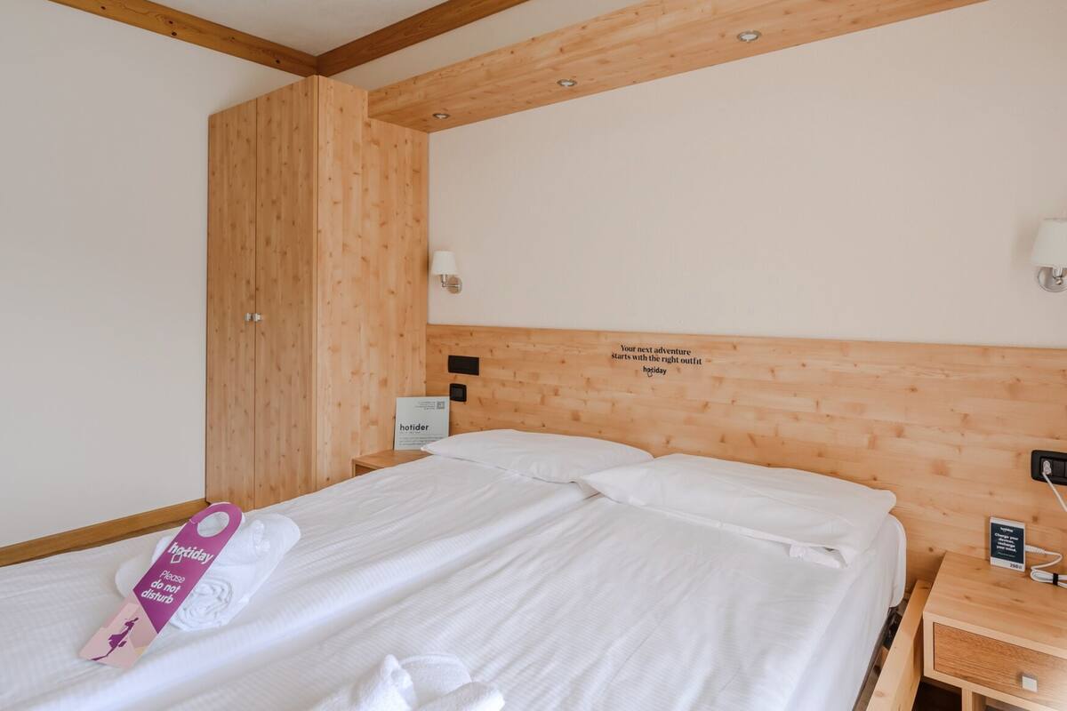 Una camera d'albergo con una testiera in legno, un letto con lenzuola bianche, un asciugamano con un cartello ‘non disturbare, vacanza’ e un cartello sul muro che recita ‘La tua prossima avventura inizia con l'abbigliamento giusto, vacanza.’