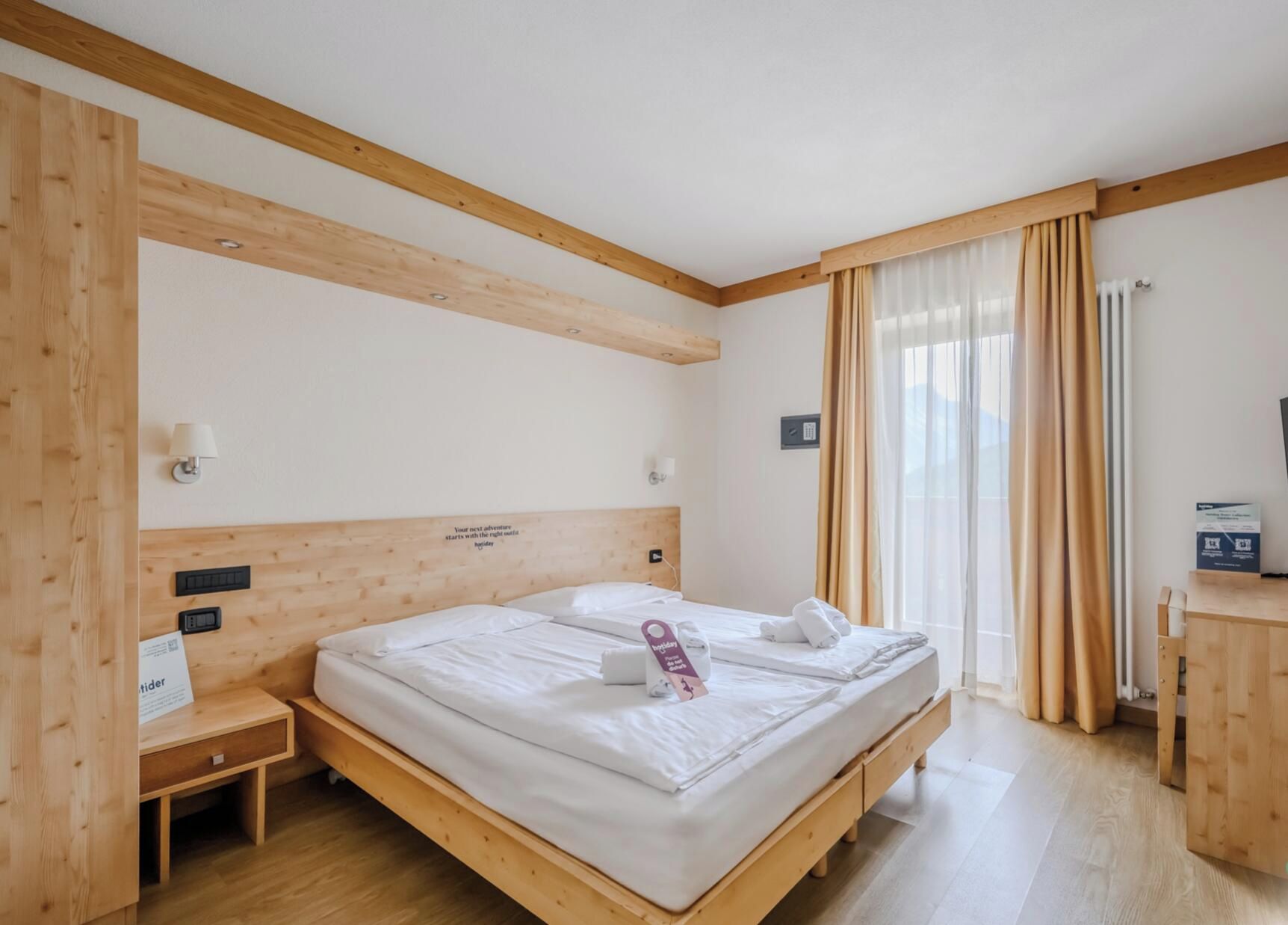 A hotel room with a wooden headboard, a bed with white linens, a bedside table, a nightstand, a window with curtains, and a small desk with a lamp.