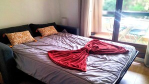 A bed with a grey bedspread and two patterned pillows, a red blanket, and a window with a view of outdoor furniture. - Flat with terrace in Sanary-sur-Mer (Sanary-sur-Mer)
