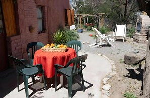 Un comedor al aire libre rústico con pared de ladrillo, una mesa con mantel rojo y sillas de plástico verde.