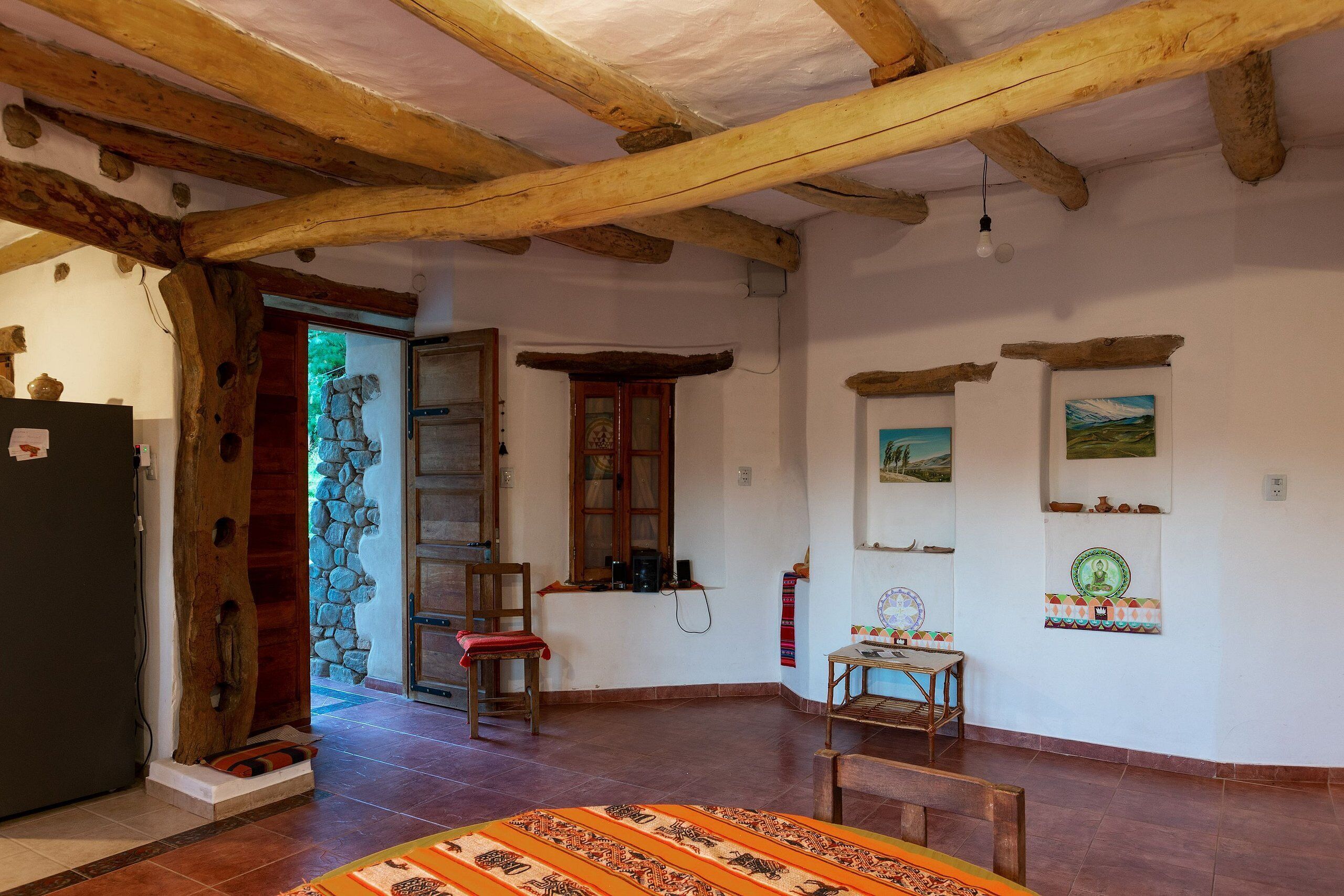 A rustic room with exposed wooden beams, a stone wall, a wooden door, a red chair, a small table, and a patterned rug.