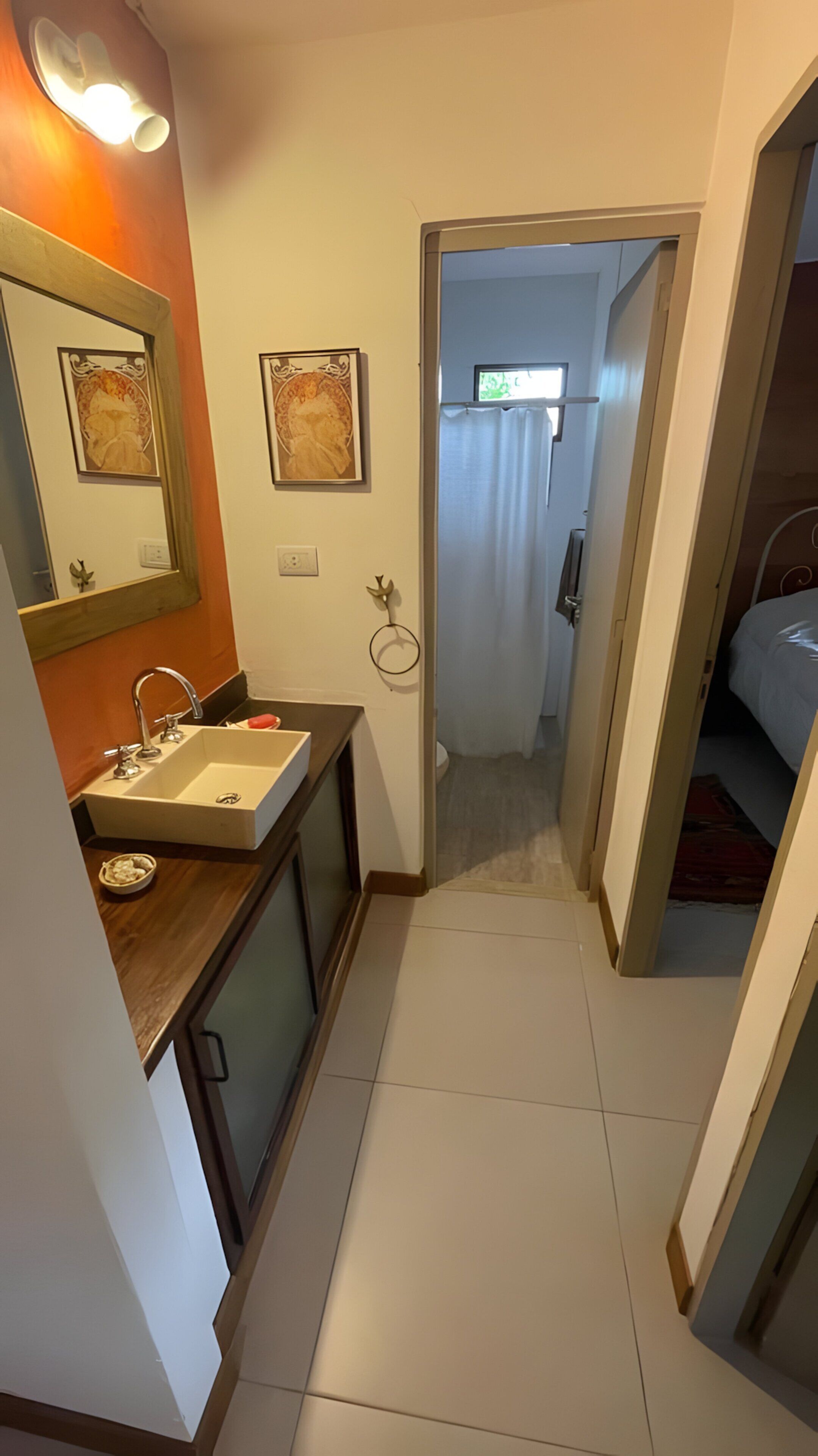 Un couloir étroit mène à une chambre à coucher avec un lit, une salle de bain avec un lavabo et un miroir, et une porte qui donne sur une autre pièce.