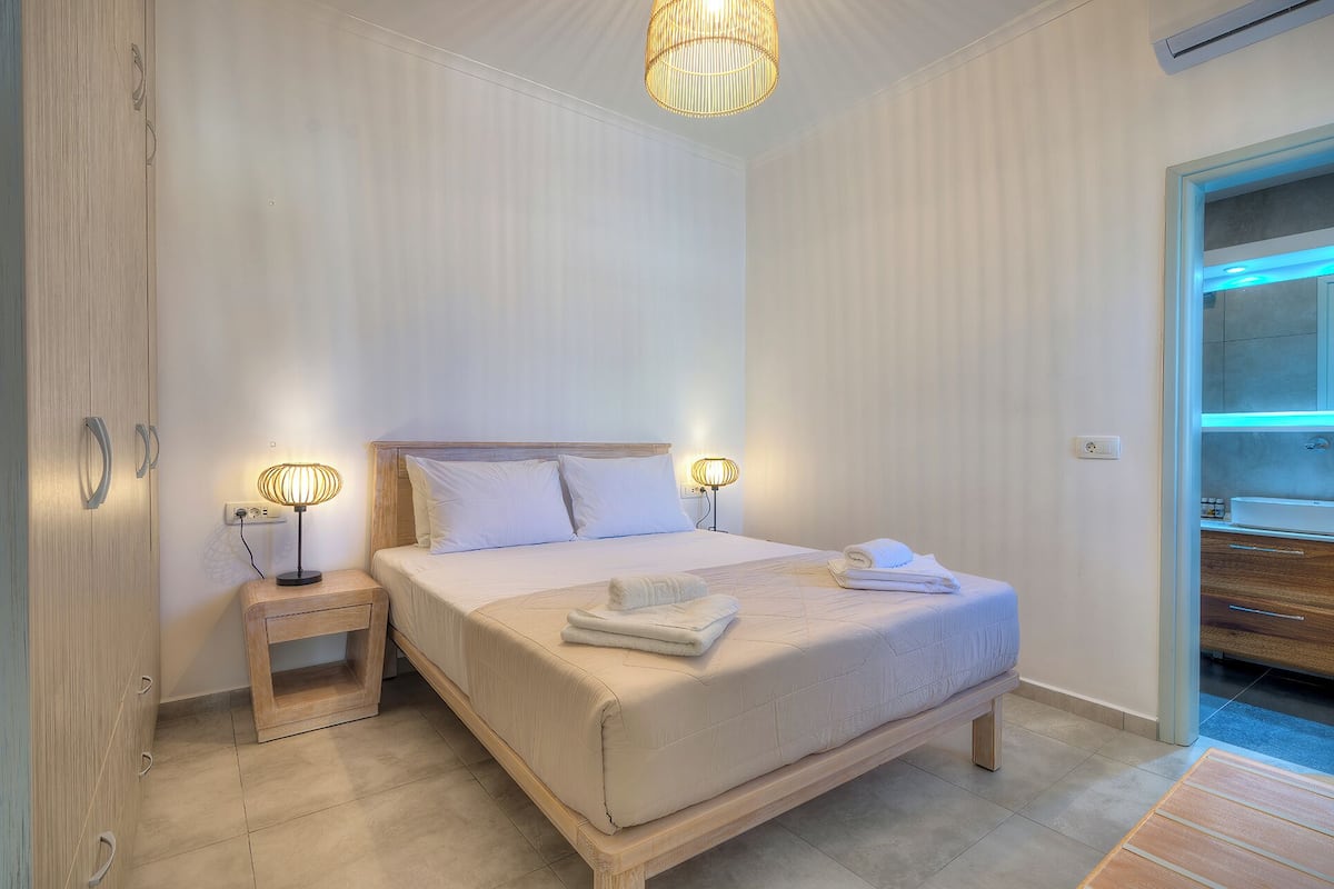 Quarto com cama, luminárias de cabeceira, mesa de cabeceira de madeira e um banheiro visível através de uma porta aberta.