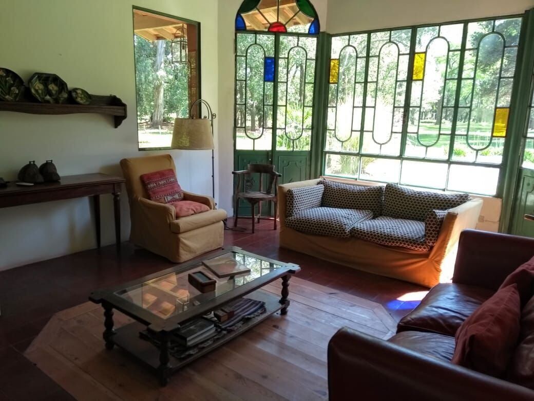 Una sala de estar acogedora con sofá, sillón y mesa de centro. Hay ventanas grandes con marcos verdes y vidrieras.