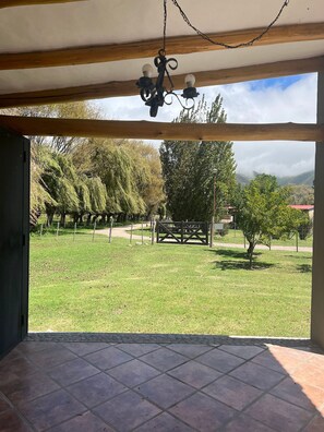 A covered patio area with a tiled floor, a hanging chandelier, and a view of a grassy landscape with trees and a distant mountain range.