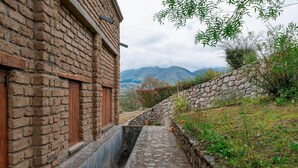 A narrow stone pathway flanked by a brick building and a stone wall, leading to a mountainous landscape.