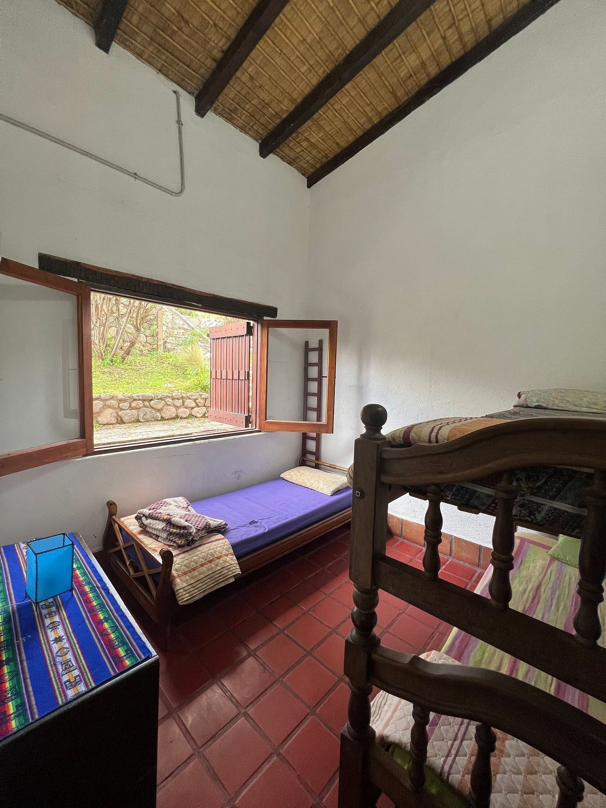 Habitació petita amb un llit individual, una finestra, una escala de fusta i una taula amb un objecte blau a sobre.