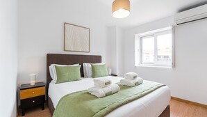 Ein ordentlich gemachtes Bett mit grün-weißer Bettwäsche, ein Nachttisch mit Lampe und ein Fenster mit Blick auf Gebäude.