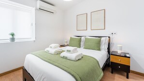 Habitación de hotel con cama, ropa de cama verde y blanca, mesita de noche con lámpara y ventana con planta.