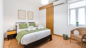 Une chambre moderne avec un lit, des tables de chevet, un plaid vert, un fauteuil en osier et une fenêtre agrémentée d’une plante.