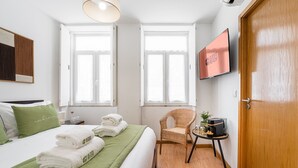 Habitación de hotel con cama, colcha verde, una silla de mimbre, una mesita con una planta y un televisor en la pared.