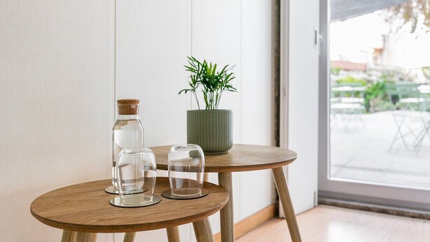 Una pequeña mesa de madera con tres vasos transparentes y una botella, una planta en maceta, y vistas a un espacio al aire libre con mesas y sillas.