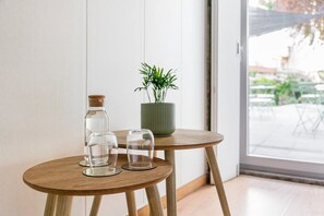 Een klein houten tafeltje met drie glazen en een fles, een potplant, en uitzicht op een buitengebied met tafels en stoelen.