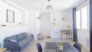 Una sala moderna con sofá, mesa de comedor y sillas. Hay una pequeña mesa con una planta, un estante montado en la pared con una planta y una lámpara en el techo.