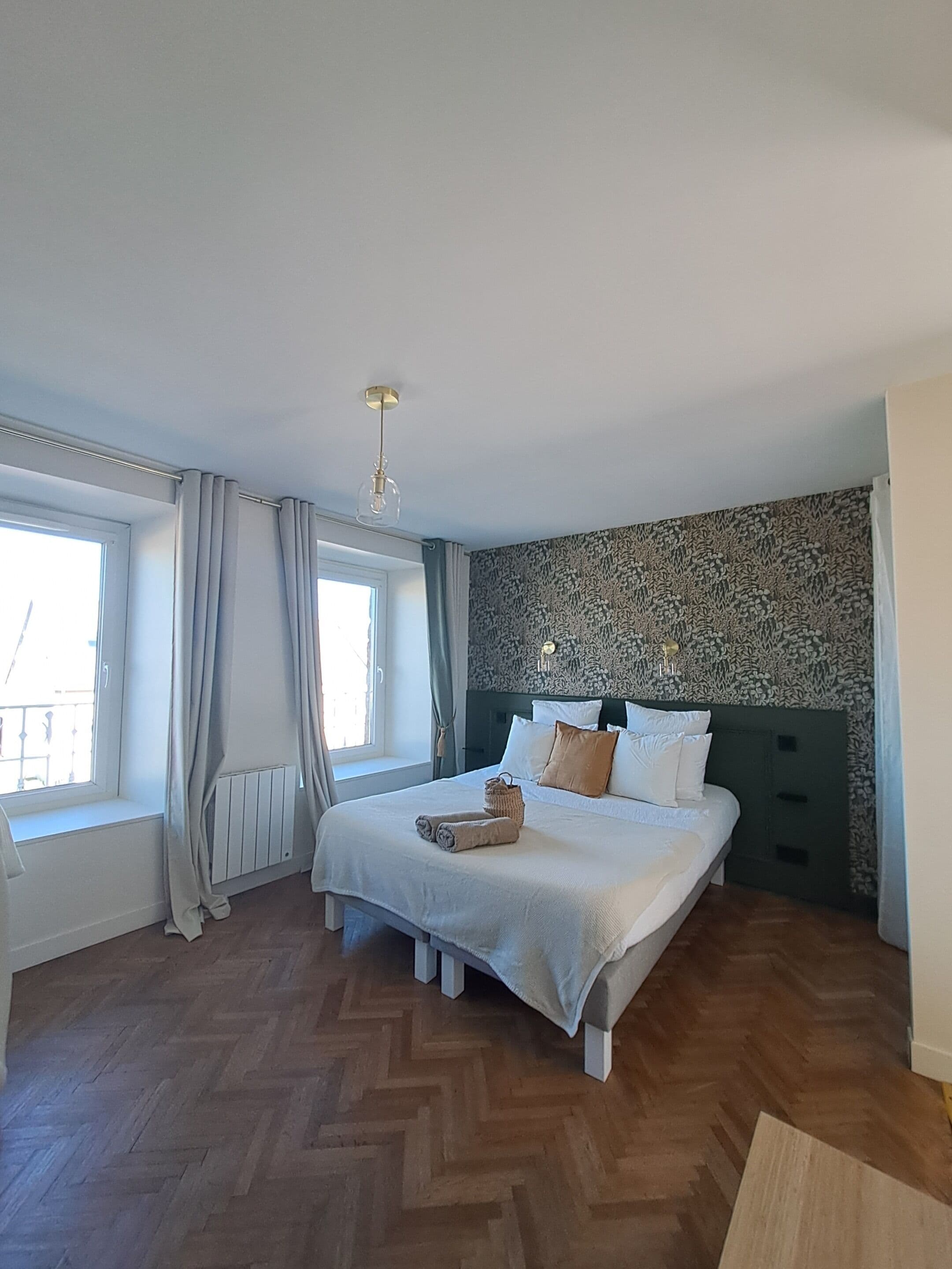 A bedroom with a large bed, a window with curtains, a patterned wall, and a wooden floor.