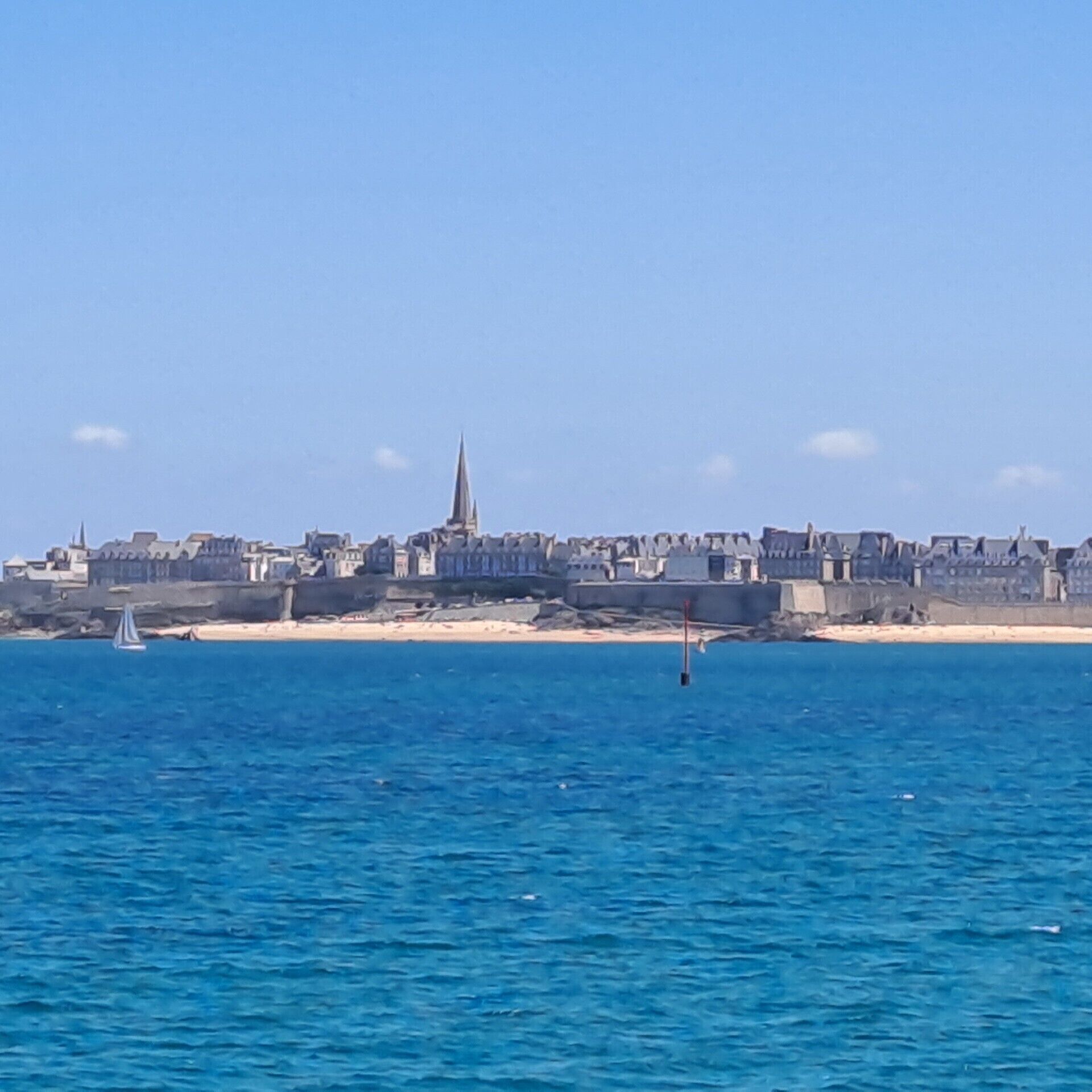 Een kuststadsgezicht met een opvallende kerktoren, zandstranden en een zeilboot op het water.