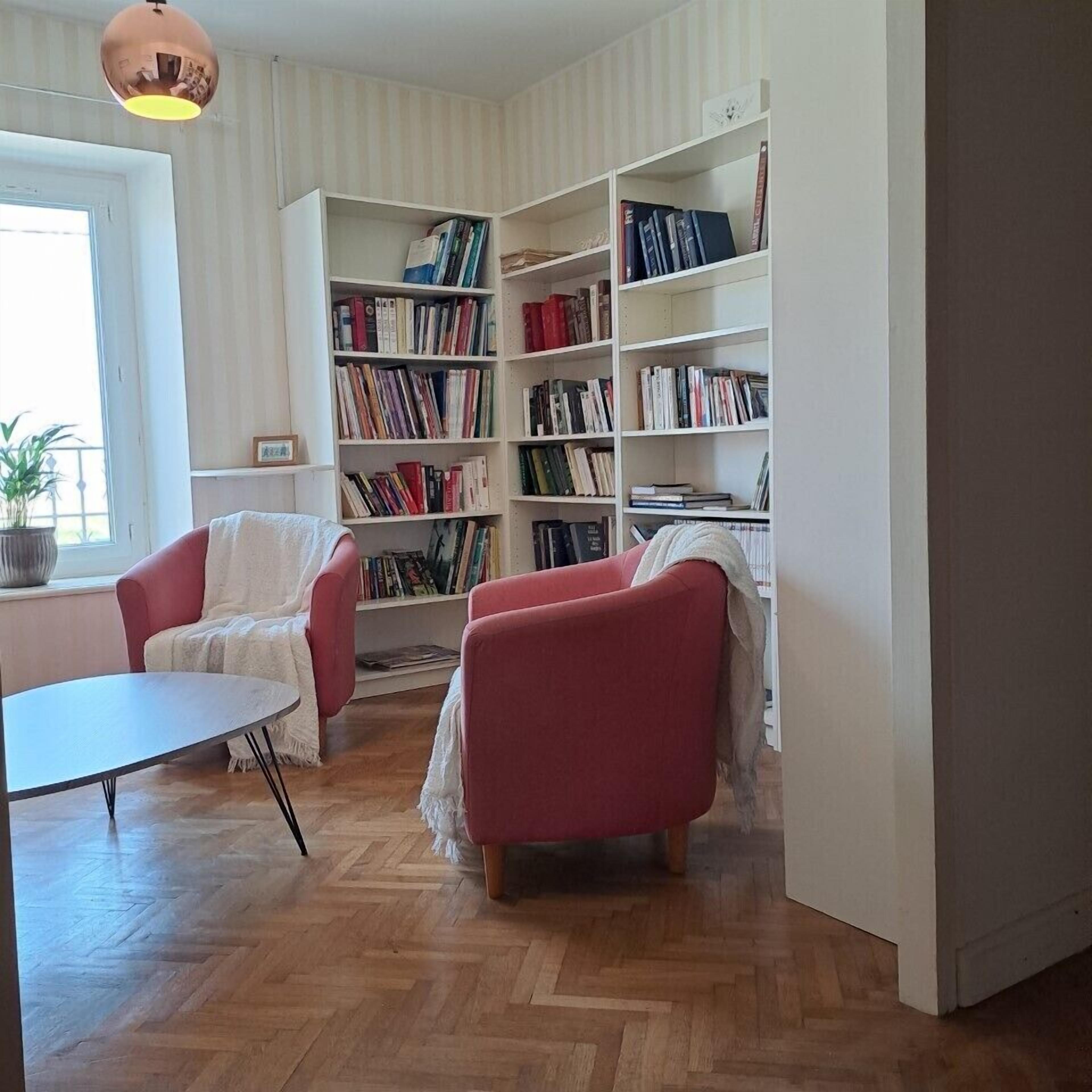 Un salon chaleureux avec un parquet en chevrons, un canapé blanc, un fauteuil rouge et une bibliothèque remplie de livres.