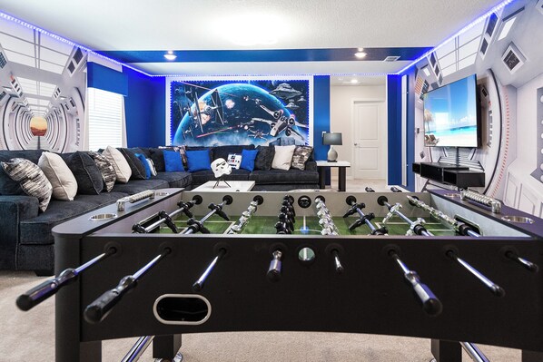 Una habitación con mesa de fútbol, sofá, pared con un mural de temática espacial y televisor.