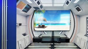 Una habitación con una pantalla grande que muestra una escena de playa, un panel de control y una zona de estar con una mesa.