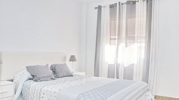 Un dormitorio con cama, dos almohadas, mesita de noche, lámpara y cortinas traslúcidas.