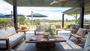A modern outdoor seating area with a sofa, chairs, and a coffee table, featuring umbrellas and a view of a golf course. - Porta Fortuna (Punta de Mita)