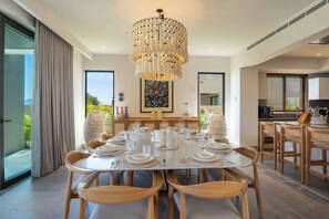 A dining area with a large round table set for six, surrounded by wooden chairs. A unique chandelier hangs above the table, and the room features large windows with a view of greenery outside.