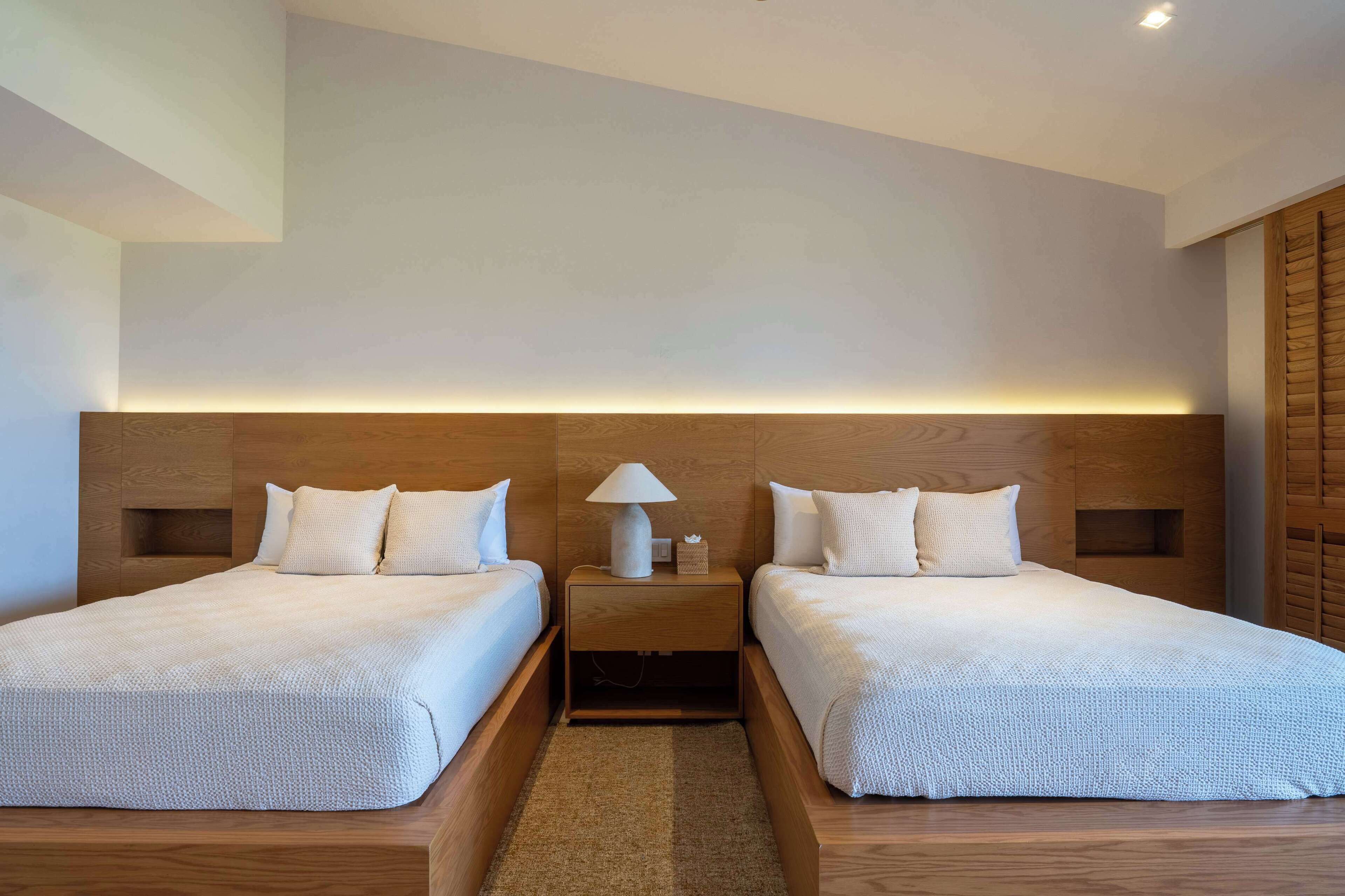 Habitación de hotel con dos camas, cabeceros de madera, una mesita de noche con lámpara y una decoración en tonos neutros.