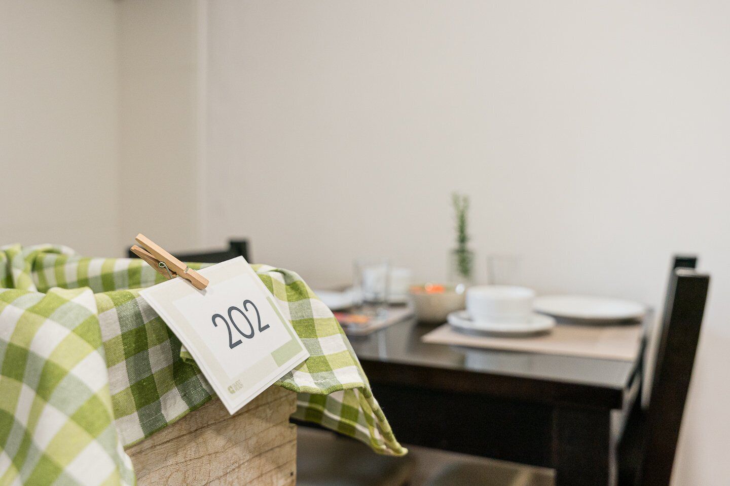 Uma área de refeições com uma mesa posta para dois, toalha quadriculada verde e branca, e o número '202' preso a um prendedor de roupa.