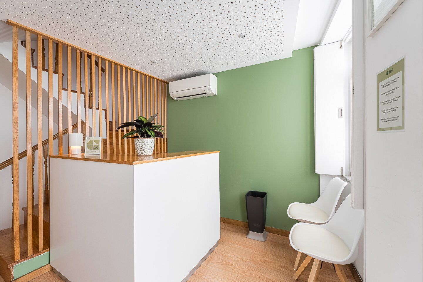 Un área de recepción con una escalera de madera, una planta en maceta, un mostrador de recepción blanco, un cubo de basura y una silla blanca.