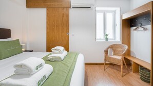 Una habitación de hotel con una cama, una colcha verde, una silla de mimbre, un armario de madera y una ventana con vistas.