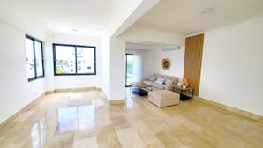 A spacious living room with a large window, a sofa, a coffee table, and a sliding door leading to a balcony.