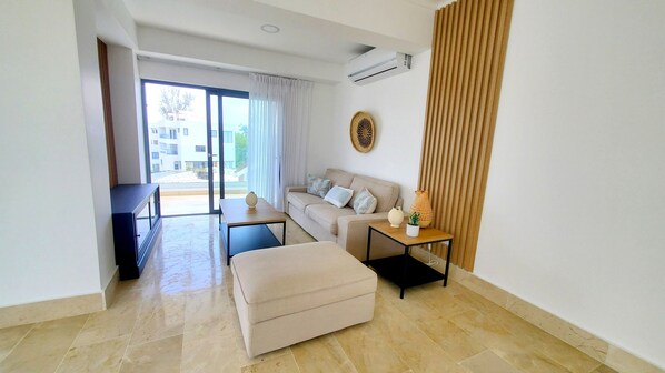 A modern living room with a sofa, ottoman, and a small table with decorative items. There is a large window with curtains, an air conditioning unit, and a view of buildings outside.