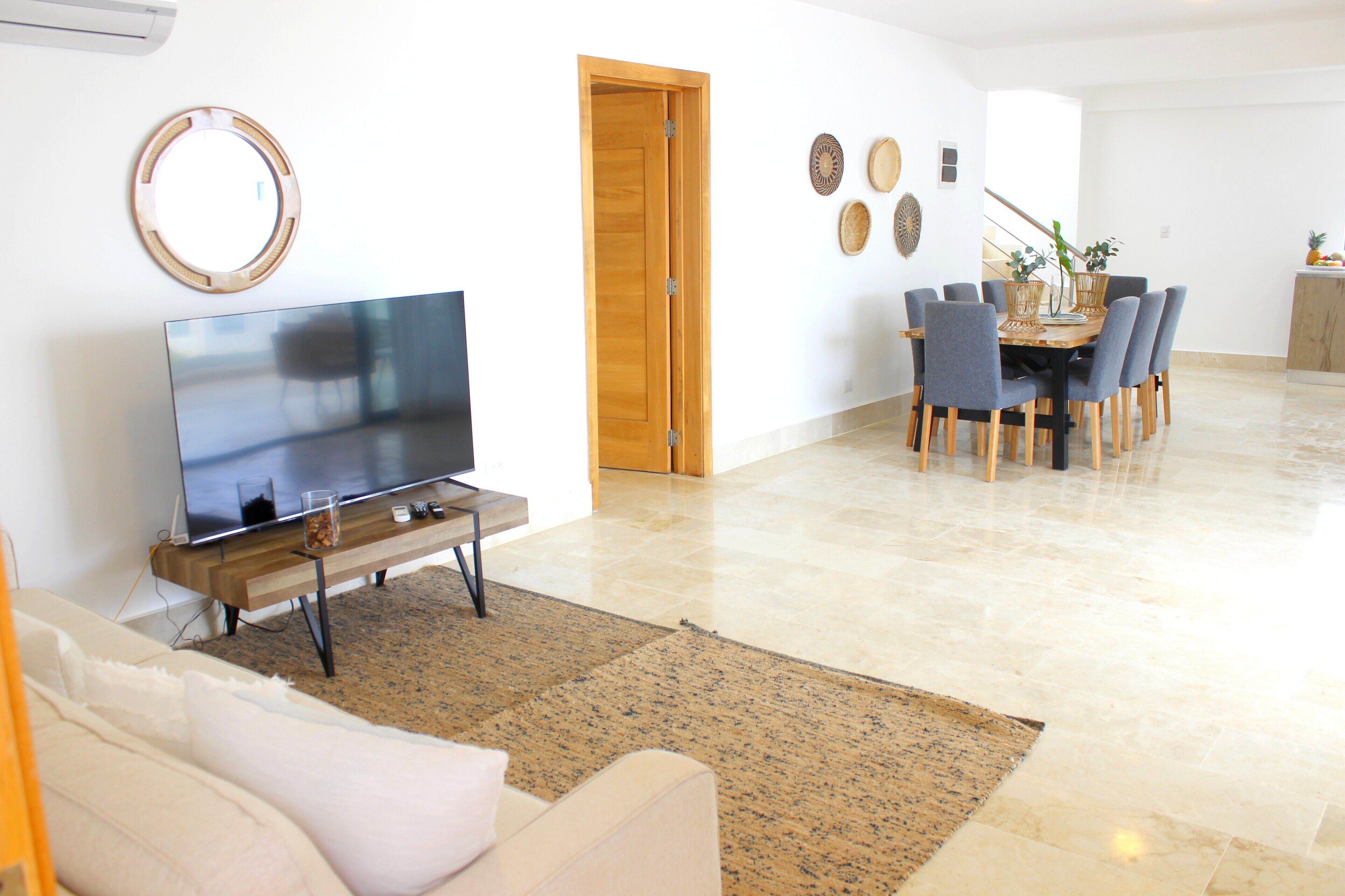 A living room with a flat-screen TV, a sofa, a dining area with chairs and a table, and a wooden door.