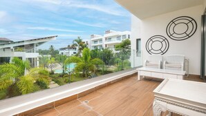 A balcony with a wooden floor, white furniture, and a view of a pool and buildings.