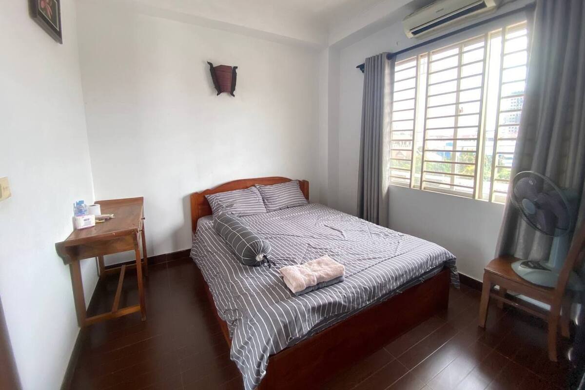 A bedroom with a bed, a wooden nightstand, a desk, a chair, and a window with curtains.