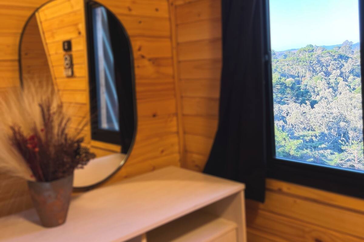 A wooden cabin interior with a round mirror, a white dresser, and a window offering a view of a forested landscape.