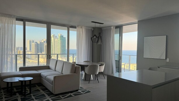 A modern living room with a sofa, a dining table with chairs, and large windows offering a city view. - 2201 (Hollywood)