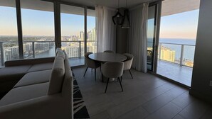 A modern living room with a sofa, dining table, and chairs. It features large windows with a city and ocean view. - 2201 (Hollywood)