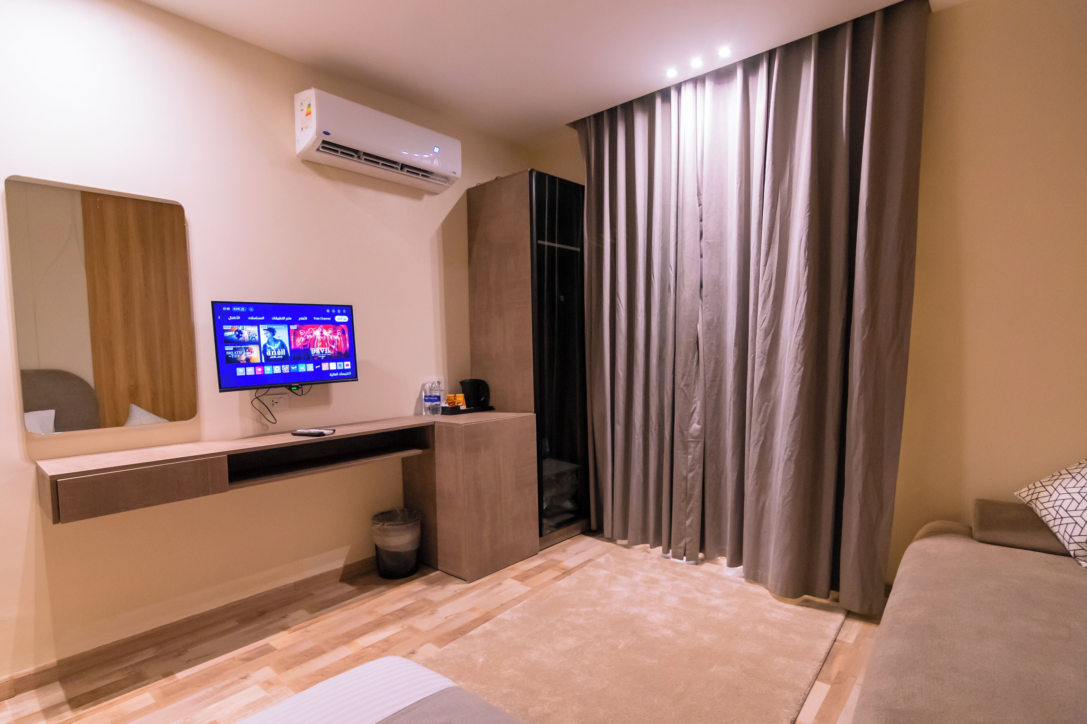 Une chambre d’hôtel équipée d’un téléviseur à écran plat fixé au mur, d’un bureau en bois, d’une poubelle, d’un canapé et d’une grande fenêtre avec des rideaux.